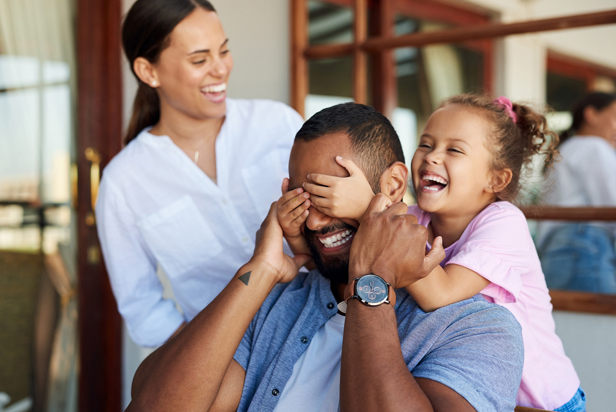 Daughter covers her fathers eyes as mother watches and laughs
