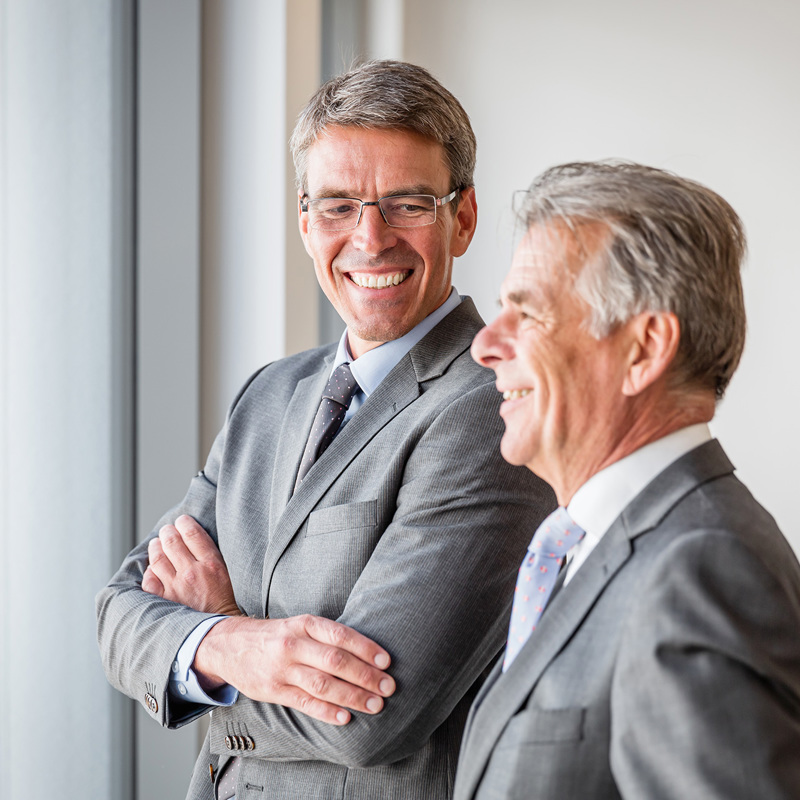 Two mature gentleman in suits looking out a window 